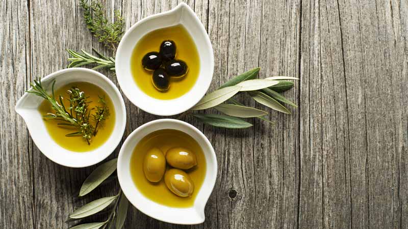3 taças de louça branca em formato de lágrima sobre uma mesa de madeira: cada taça tem azeite, temperos e azeitonas - alusão ao azeite